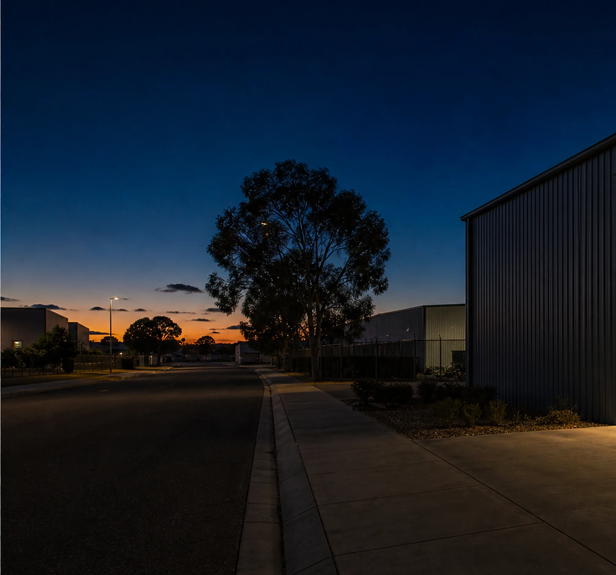 A quiet industrial street at dusk, a gum tree and the dark wall of a shed